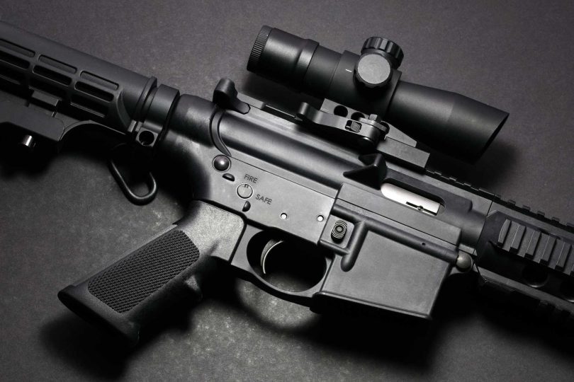 Close-up of an AR-15 receiver area showing the upper and lower receiver, trigger, magazine well, and optic mounted on the rail