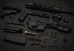 Disassembled AR-15 rifle parts arranged on a dark workbench, including stock, grip, upper and lower receivers, barrel assembly, magazine, and small components