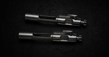 Two metallic bolt carrier groups placed parallel on a dark textured surface with soft directional lighting