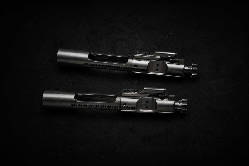 Two metallic bolt carrier groups placed parallel on a dark textured surface with soft directional lighting