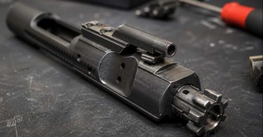 Close-up of a metal bolt carrier group resting on a dark workbench with tools blurred in the background