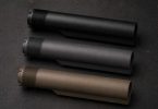 Three AR-style buffer tubes in black, gray, and tan finishes arranged diagonally on a dark workbench surface