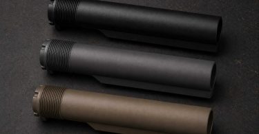 Three AR-style buffer tubes in black, gray, and tan finishes arranged diagonally on a dark workbench surface
