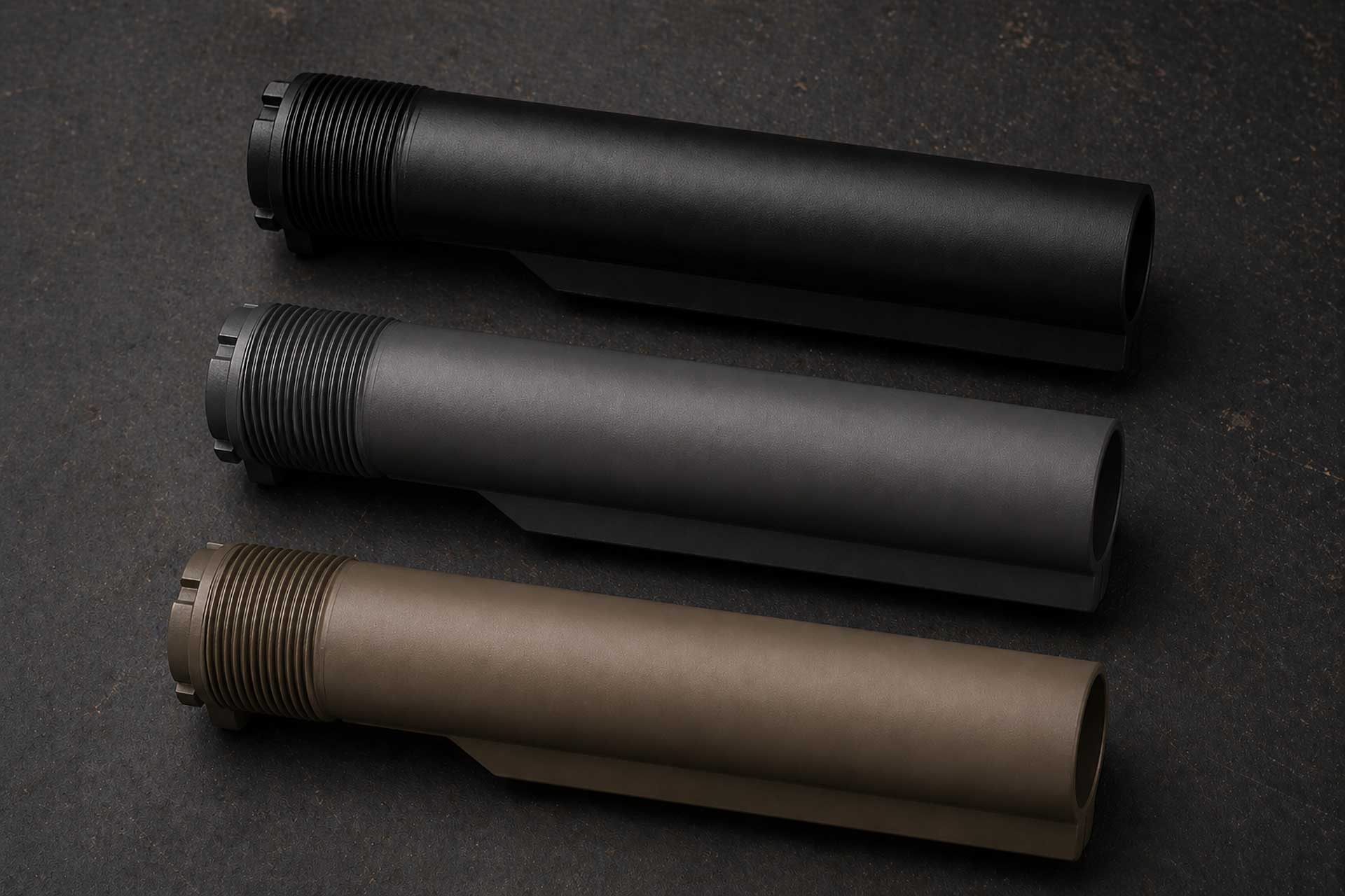 Three AR-style buffer tubes in black, gray, and tan finishes arranged diagonally on a dark workbench surface