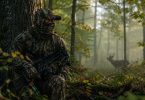 Hunter wearing woodland camouflage while sitting against a tree in a misty forest with a deer visible in the background.