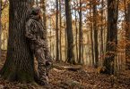 Hunter wearing camouflage while standing beside a large oak tree in an autumn hardwood forest.