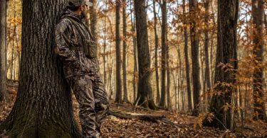Hunter wearing camouflage while standing beside a large oak tree in an autumn hardwood forest.