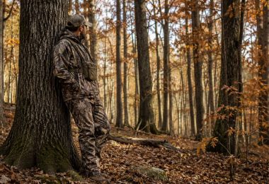 Hunter wearing camouflage while standing beside a large oak tree in an autumn hardwood forest.