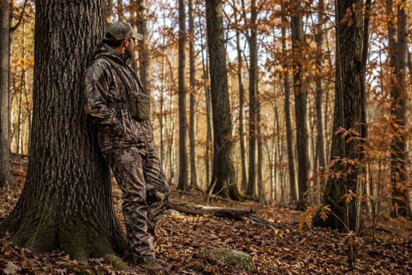 Hunter wearing camouflage while standing beside a large oak tree in an autumn hardwood forest.