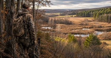 Hunter in camouflage overlooking a landscape with forest, wetlands, and open fields.