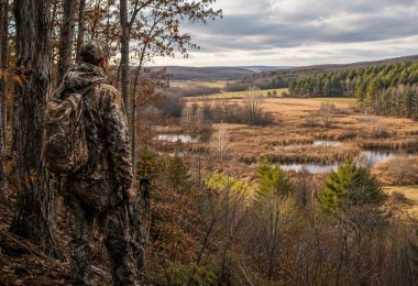 Hunter in camouflage overlooking a landscape with forest, wetlands, and open fields.