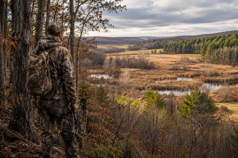 Hunter in camouflage overlooking a landscape with forest, wetlands, and open fields.