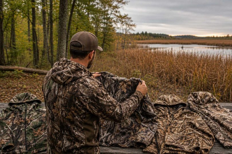 Hunter comparing several camouflage jackets beside a marsh and woodland landscape